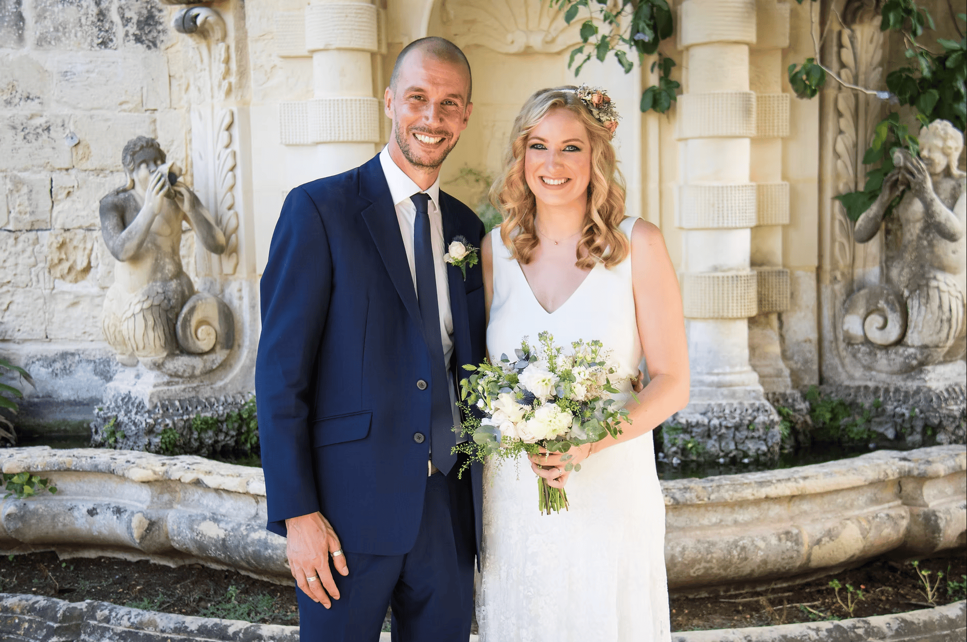 Smiling bride and groom posing together by an ornate stone fountain